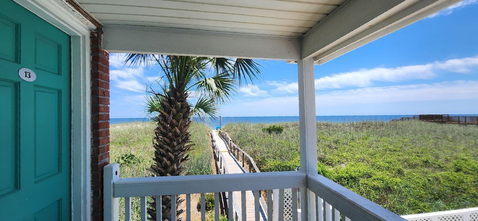 View from a balcony showing a wooden walkway leading through beach grass to the ocean on a sunny day.