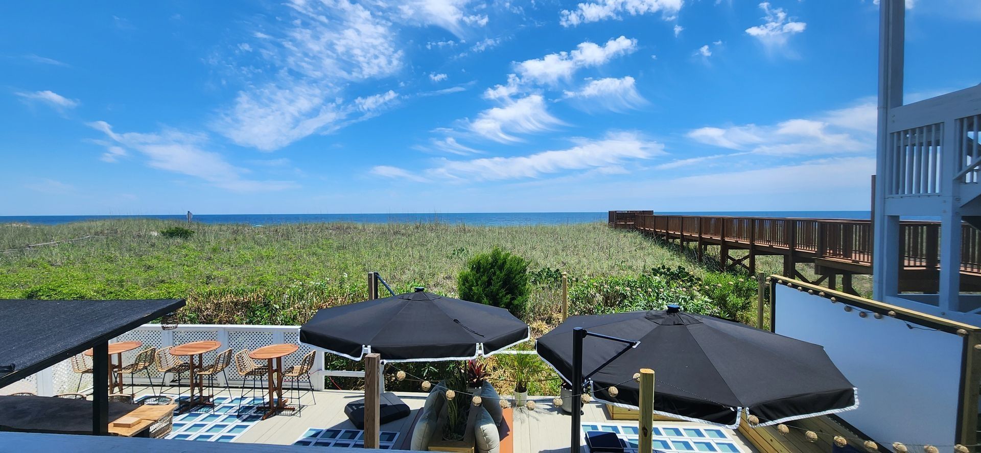 A sunny outdoor patio with black umbrellas overlooks a grassy dune area leading to a wooden boardwalk and the ocean.
