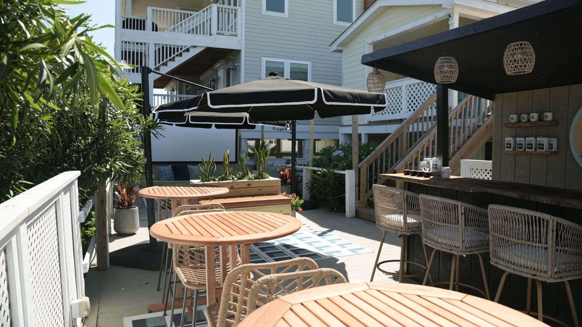 An outdoor patio seating area with round wooden tables, wicker chairs, and black umbrellas next to a bar.