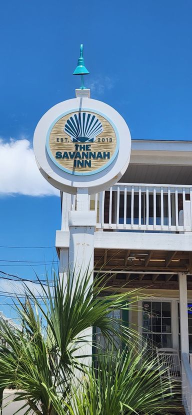 A round, white sign for The Savannah Inn with a blue seashell logo, mounted on a post against a bright blue sky.