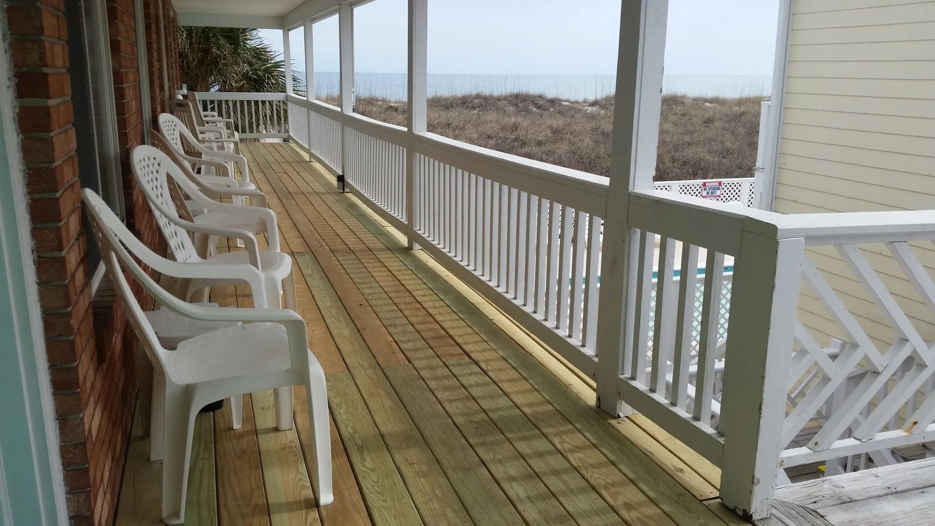 A wooden porch with a row of white plastic chairs overlooking a sandy beach and the ocean under an overcast sky.