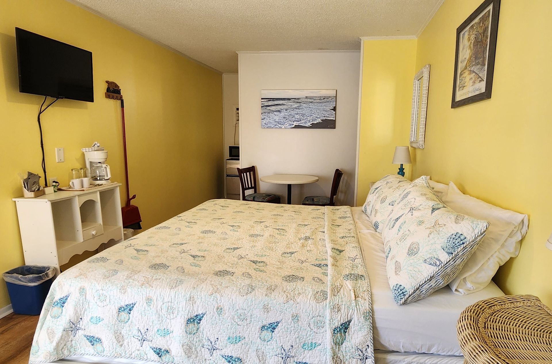 A hotel room with yellow walls, a king bed with shell-patterned bedding, a TV, a coffee station, and a small dining table.