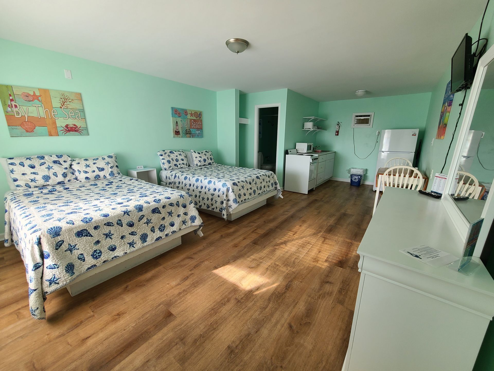 A brightly lit, mint-green hotel room featuring two beds with blue and white patterned bedding and light wood flooring.