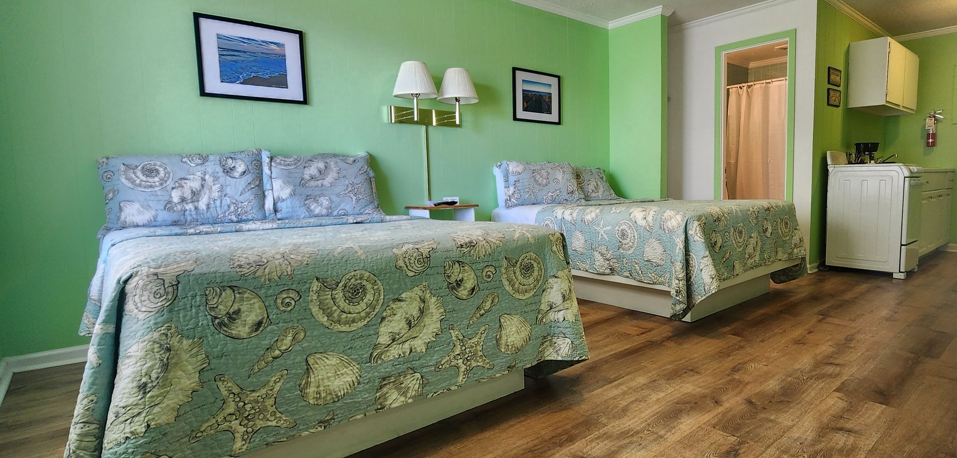 A hotel room with light green walls, two beds with seashell-patterned bedding, hardwood floors, and an adjacent bathroom.