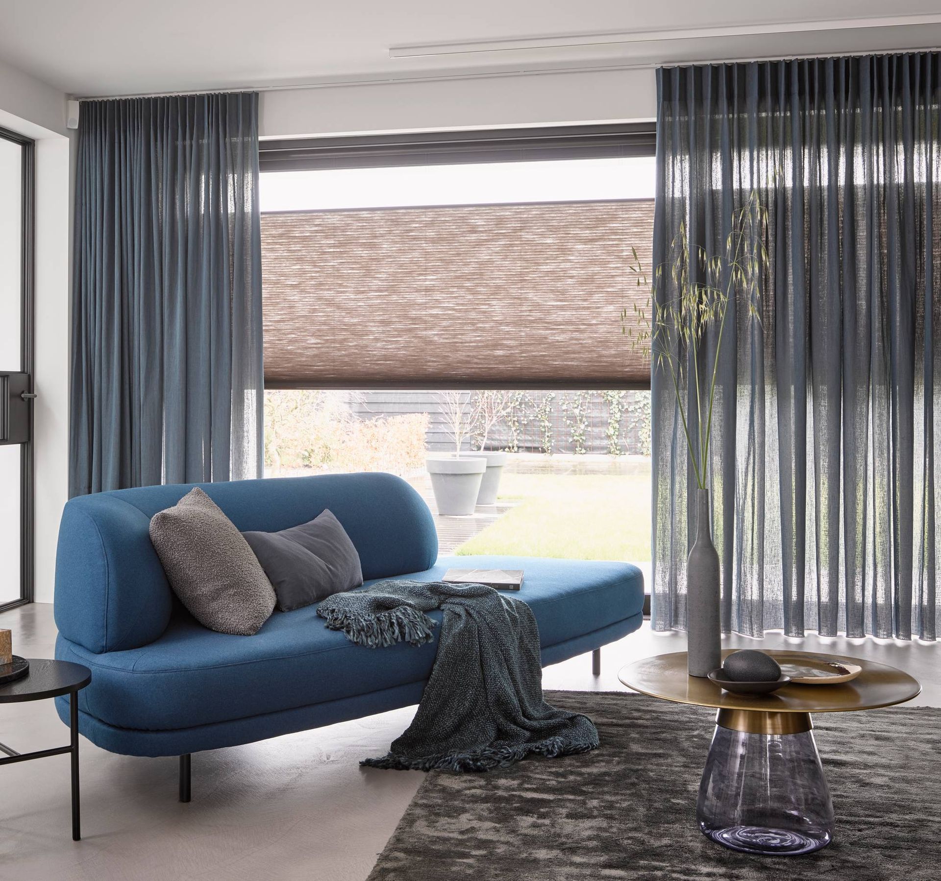 Moderne woonkamer met een blauwe chaise longue, grijze kussens, vitrage en een geweven rolgordijn voor het raam