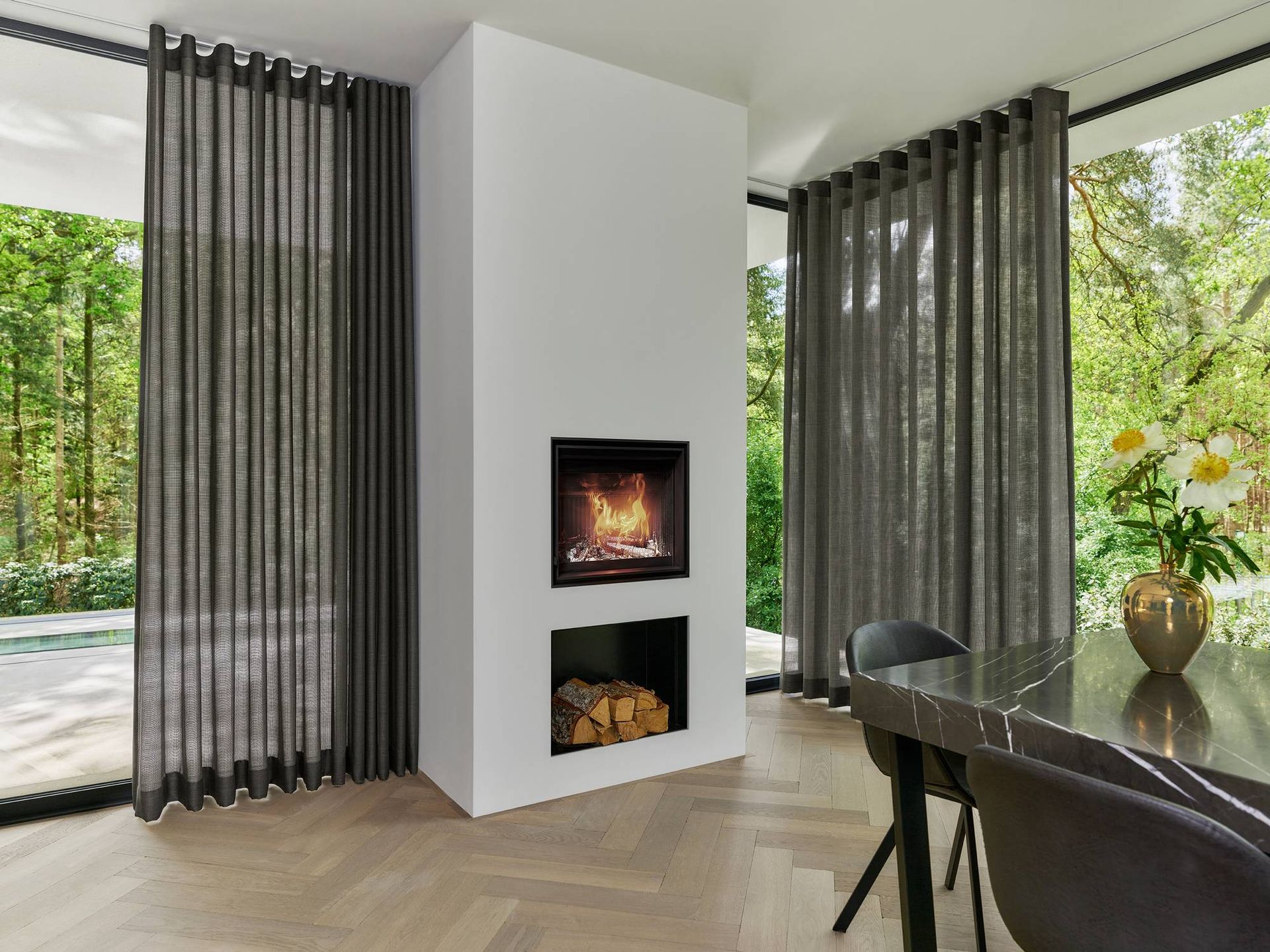 Moderne woonkamer met kamerhoge ramen, een centrale open haard en donkere gordijnen naast een eettafel.