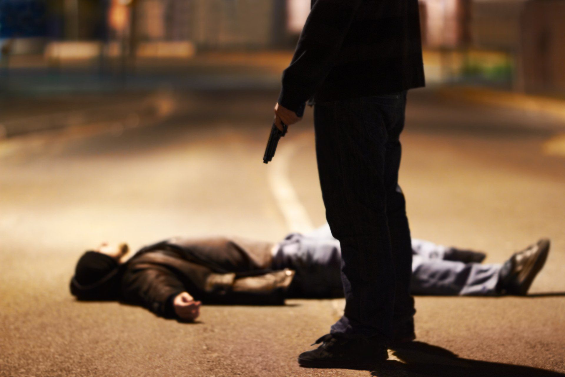 Man with gun standing over person lying on the ground in a dark street.