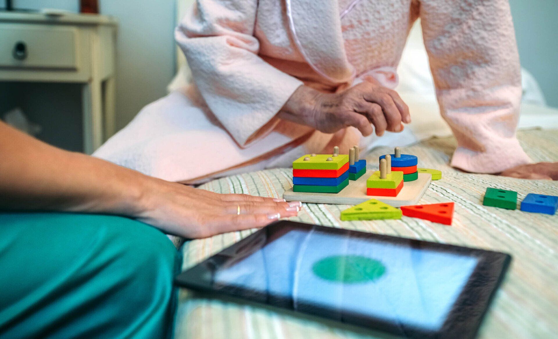 Woman playing a shape sorting game with a caregiver, tablet in foreground, on a bed.