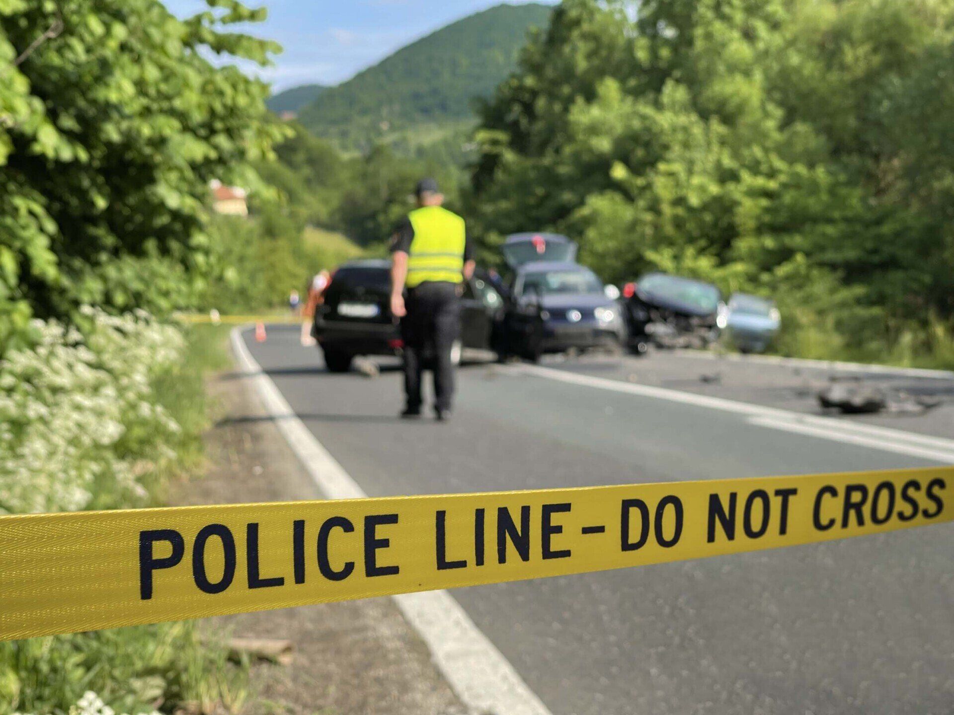 Police officer at a car accident scene. Yellow police tape across road, damaged cars, green trees, mountain in the distance.