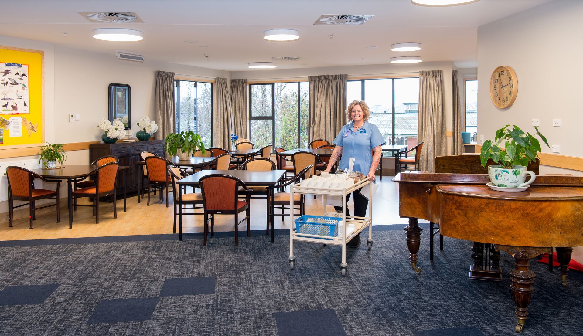 Woman pushing a cart in a sunny room with tables, chairs, and a piano.