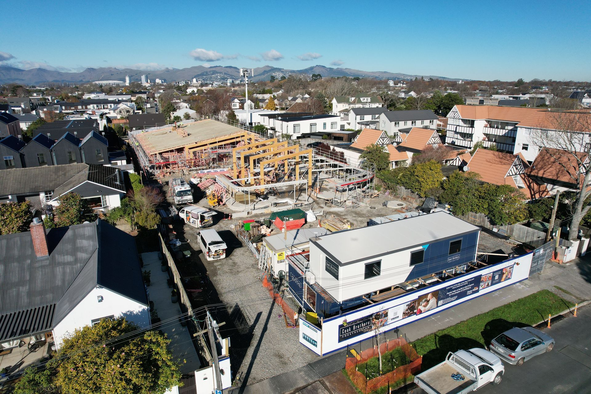 Construction site with wooden frame of a building, surrounded by houses, street, and construction trailers under a blue sky.