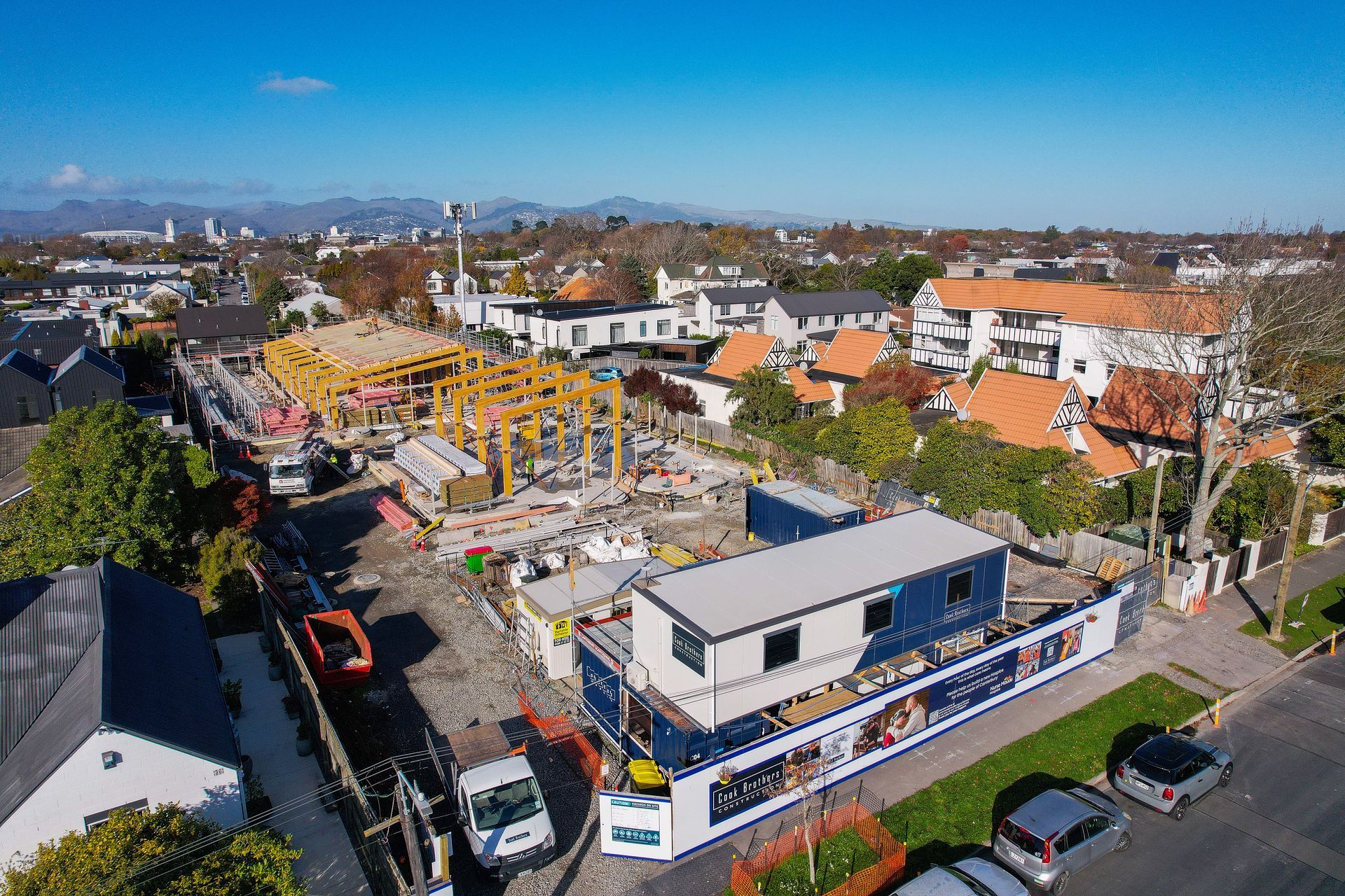 Construction site with wood framing, white structures, blue temporary buildings, and residential houses in the background.