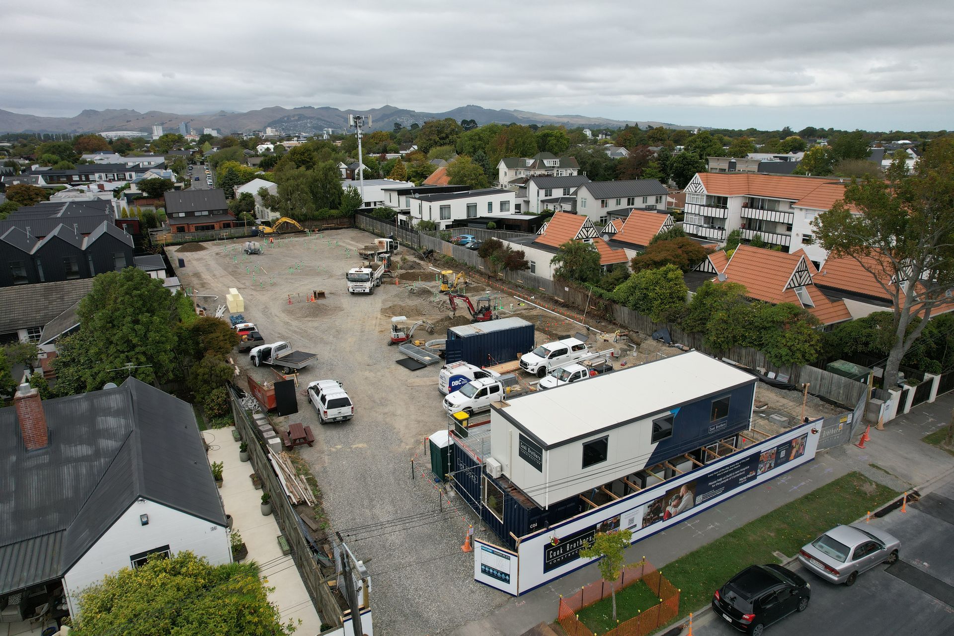 Construction site with buildings and vehicles. White and blue mobile office in foreground. Overcast sky.