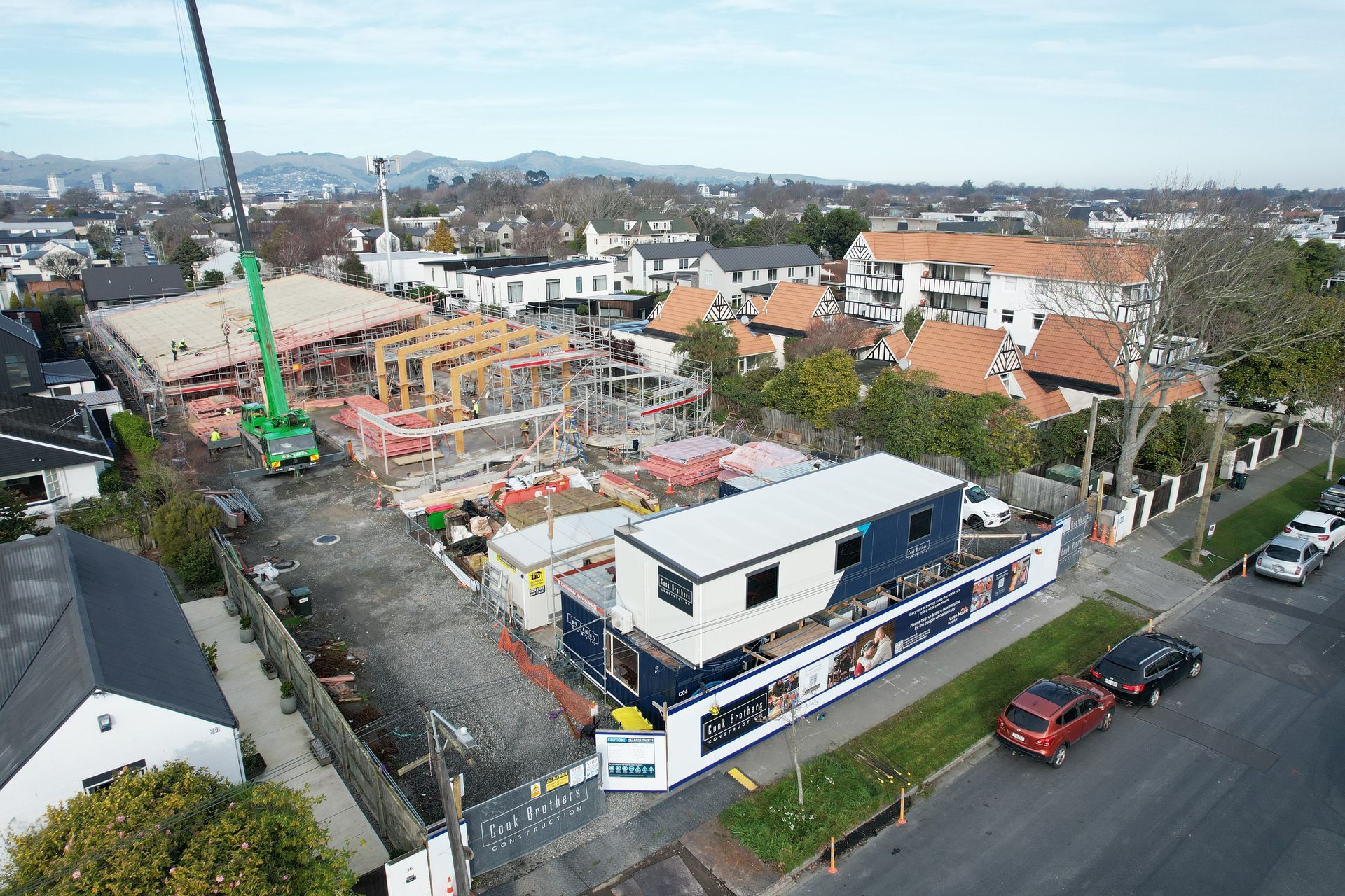 Construction site with a crane lifting materials, near houses and a street with parked cars.
