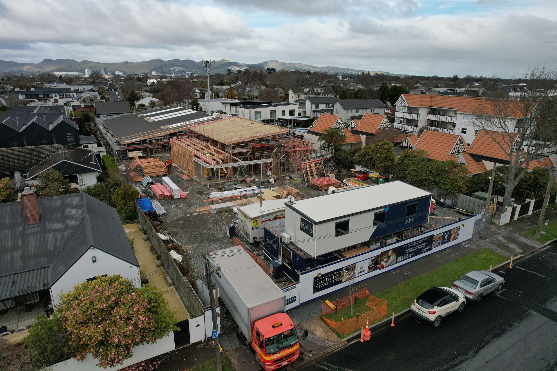 Construction site with partially built building; portable offices, truck, houses, and trees. Cloudy sky.