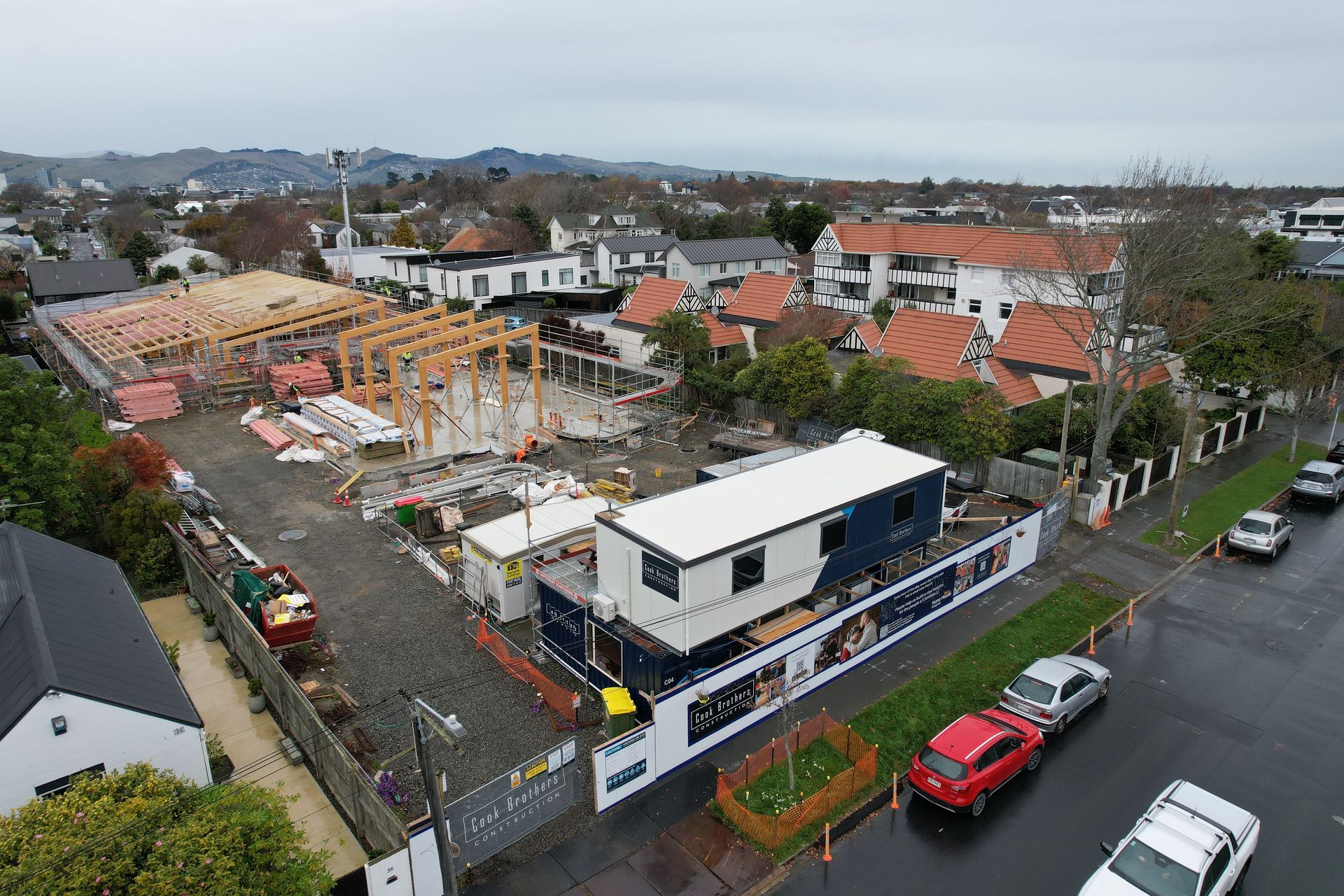 Construction site with wooden framing, a trailer, and surrounding buildings on a cloudy day.