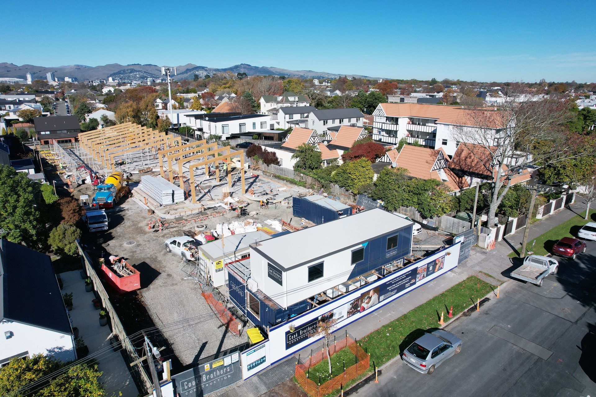 Construction site with wooden framework, container offices, and surrounding houses under a blue sky.
