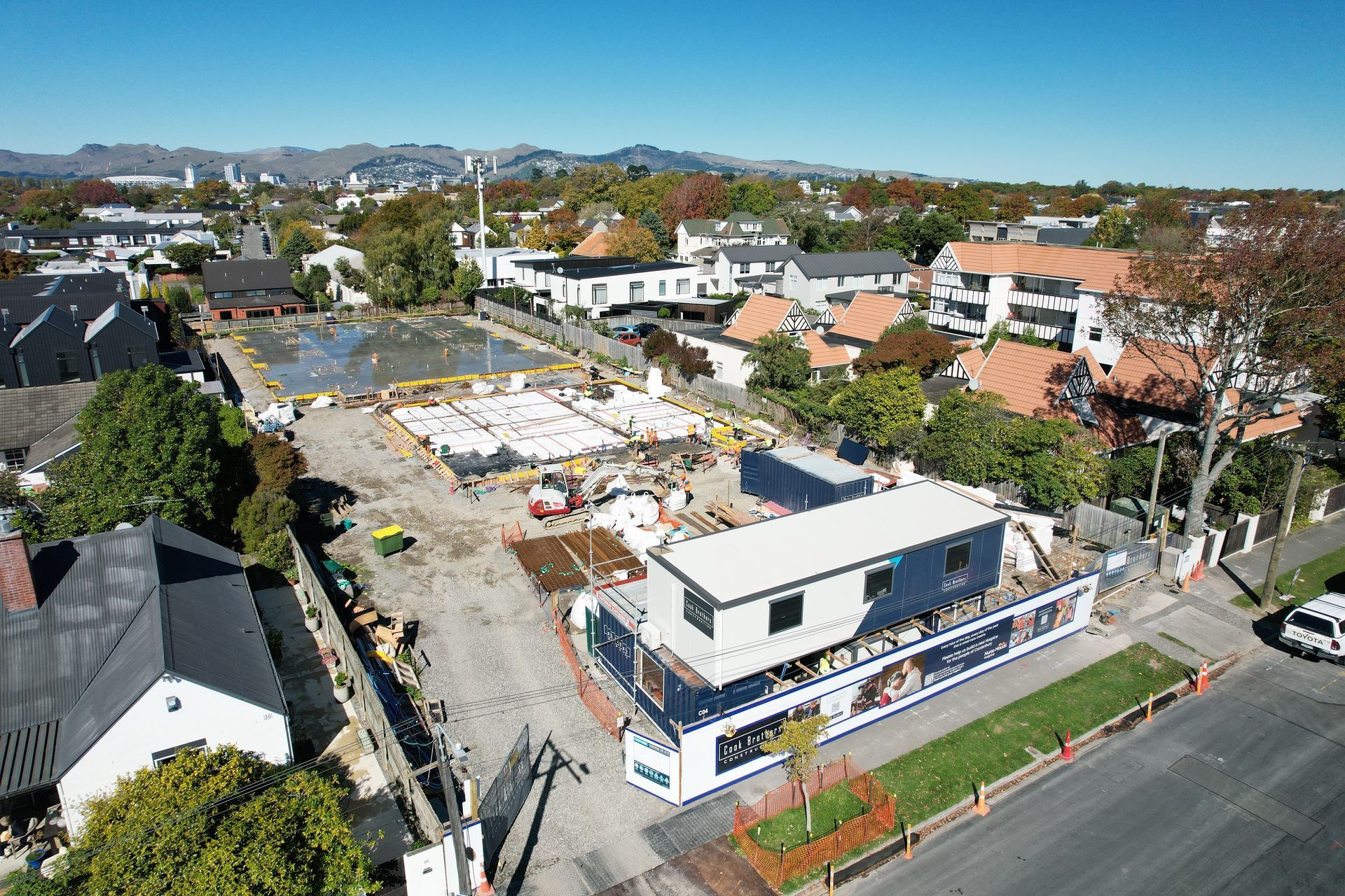 Construction site with modular buildings, concrete foundations, and residential neighborhood under a blue sky.
