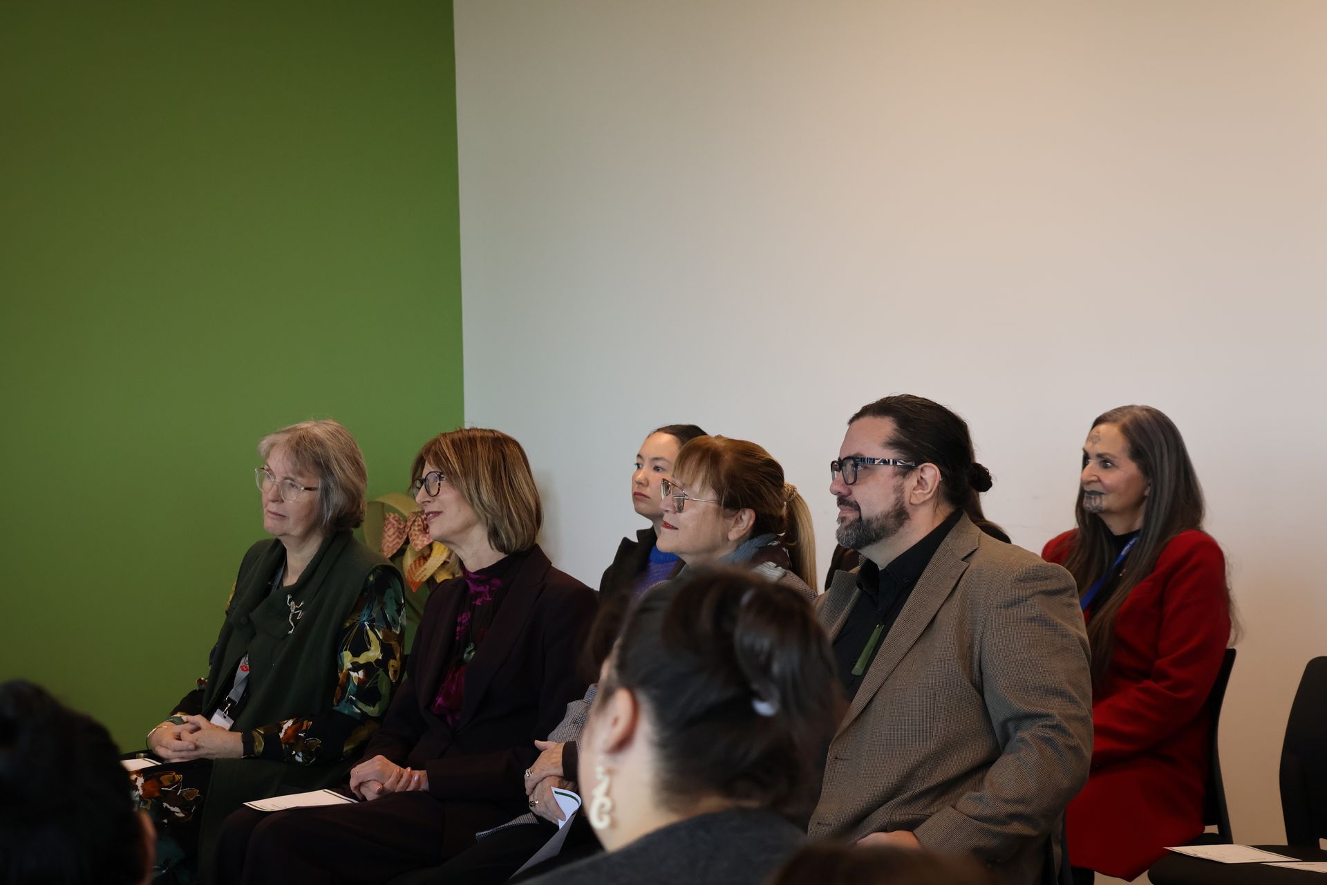 Audience seated in a conference room, listening attentively against a green and white wall.