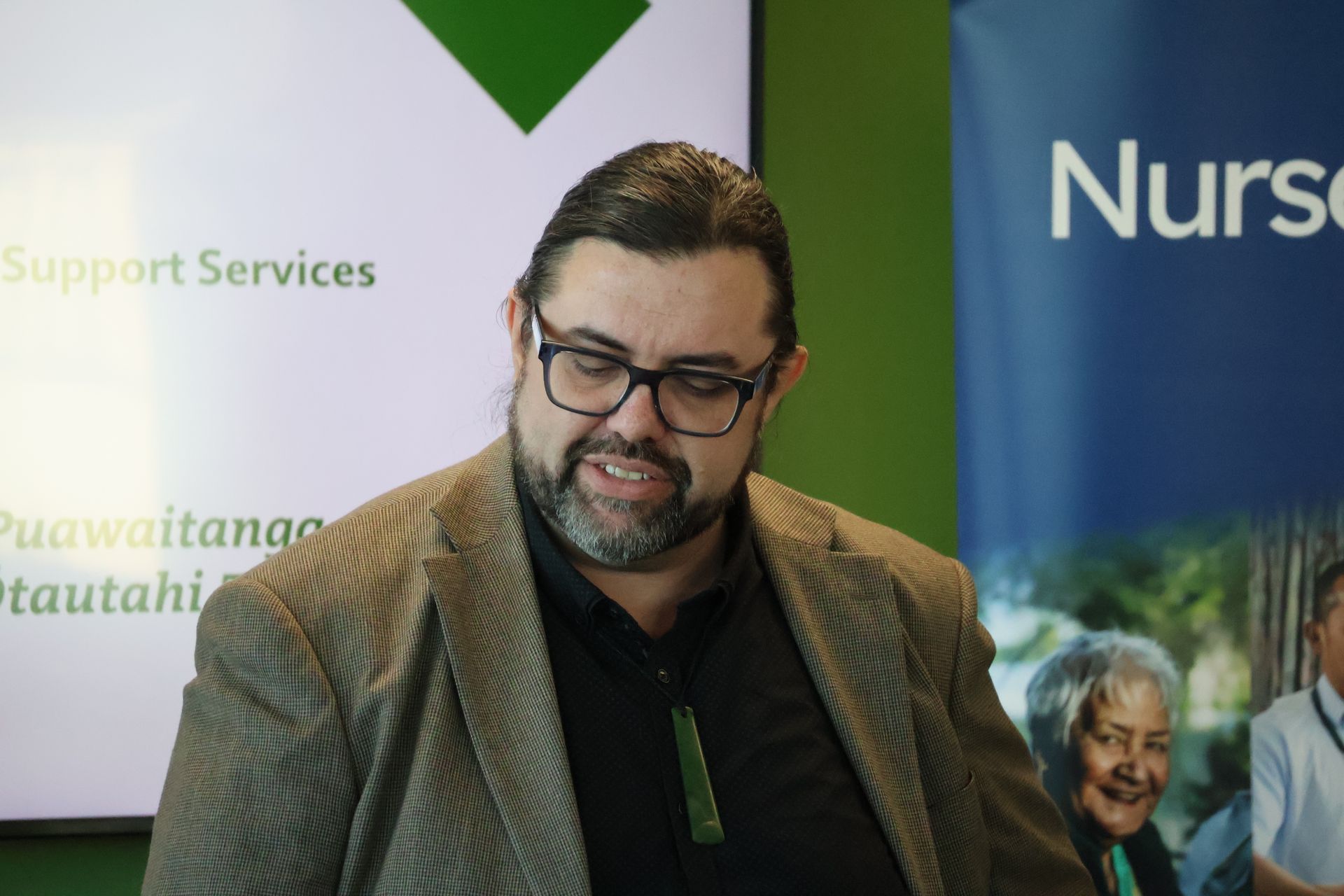 Man in glasses and brown jacket speaking at a healthcare event with support services banners behind him