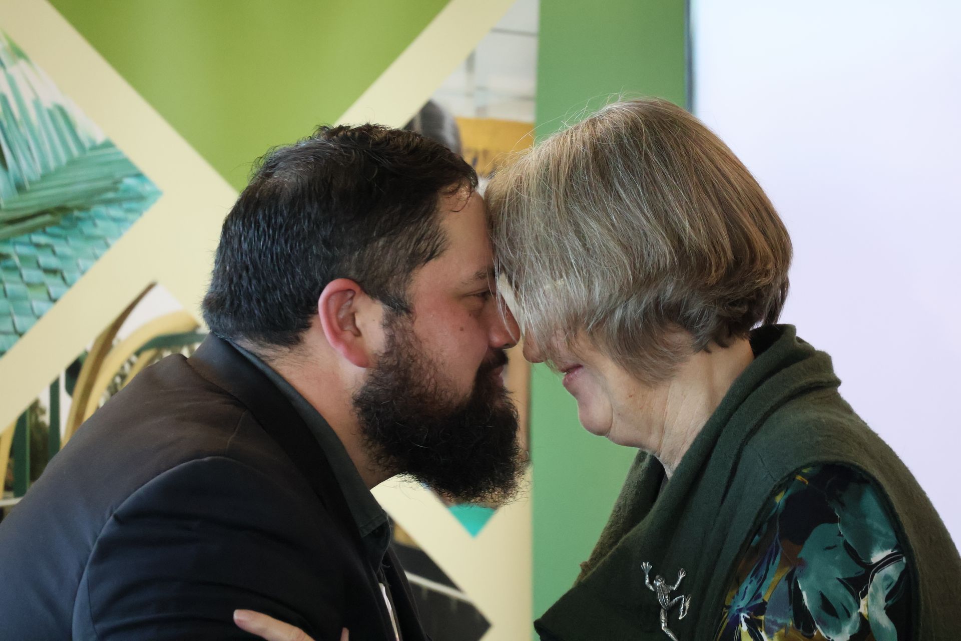 Two people lean in forehead to forehead, sharing an intimate moment in front of a green and white wall.