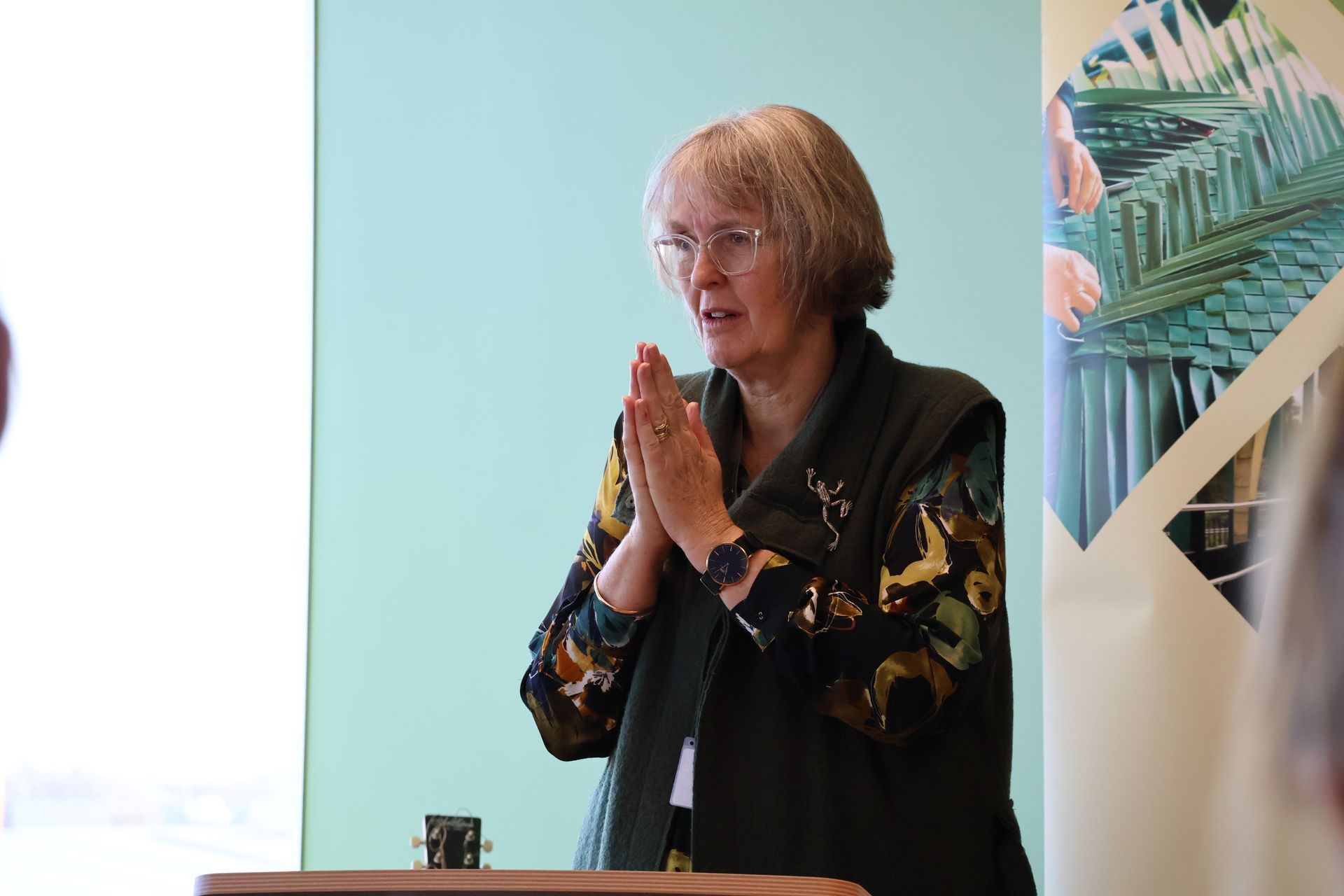 Woman speaking at a podium, hands clasped, against a light blue wall with a banner