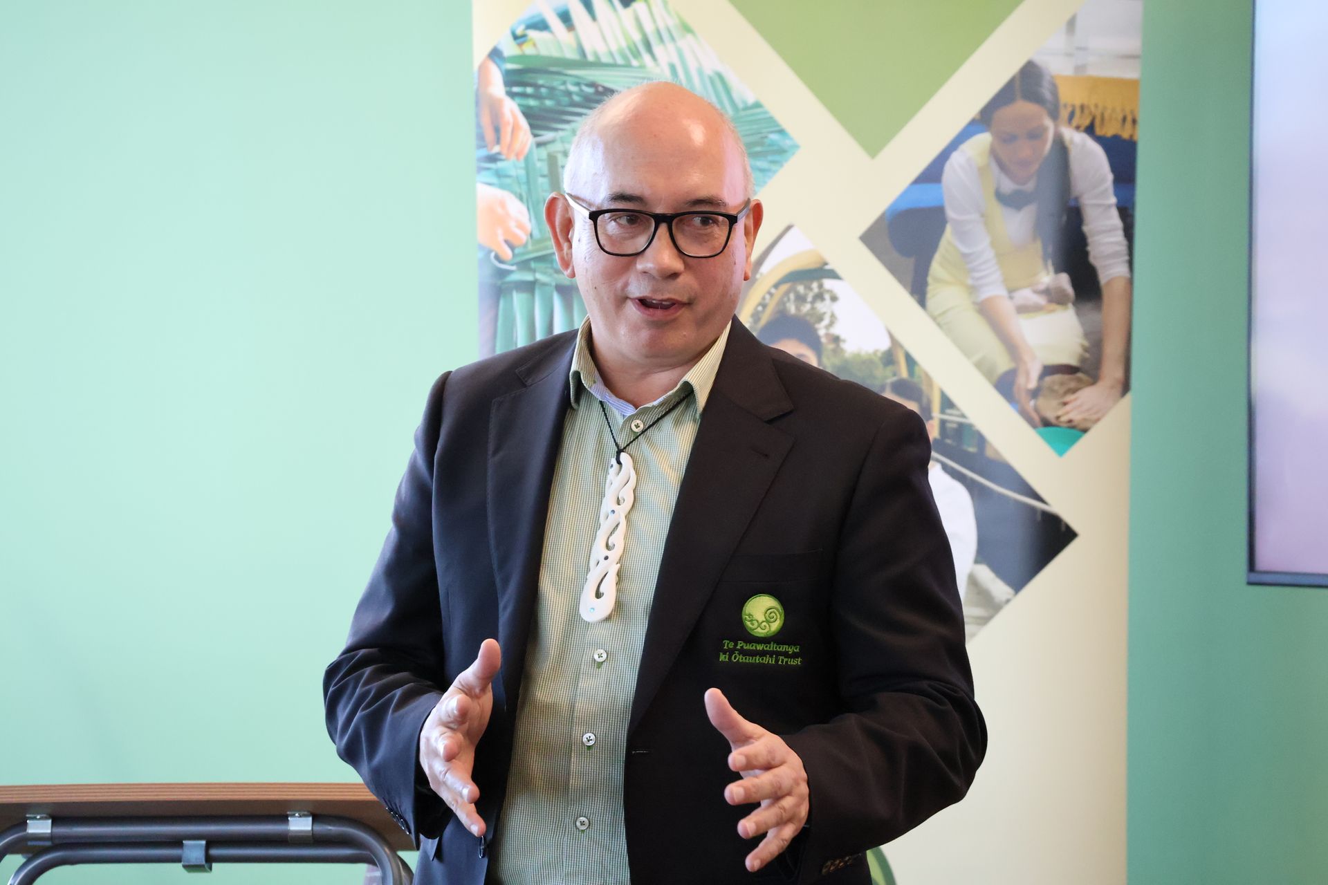 Man in a dark blazer speaking indoors beside a green wall and geometric poster
