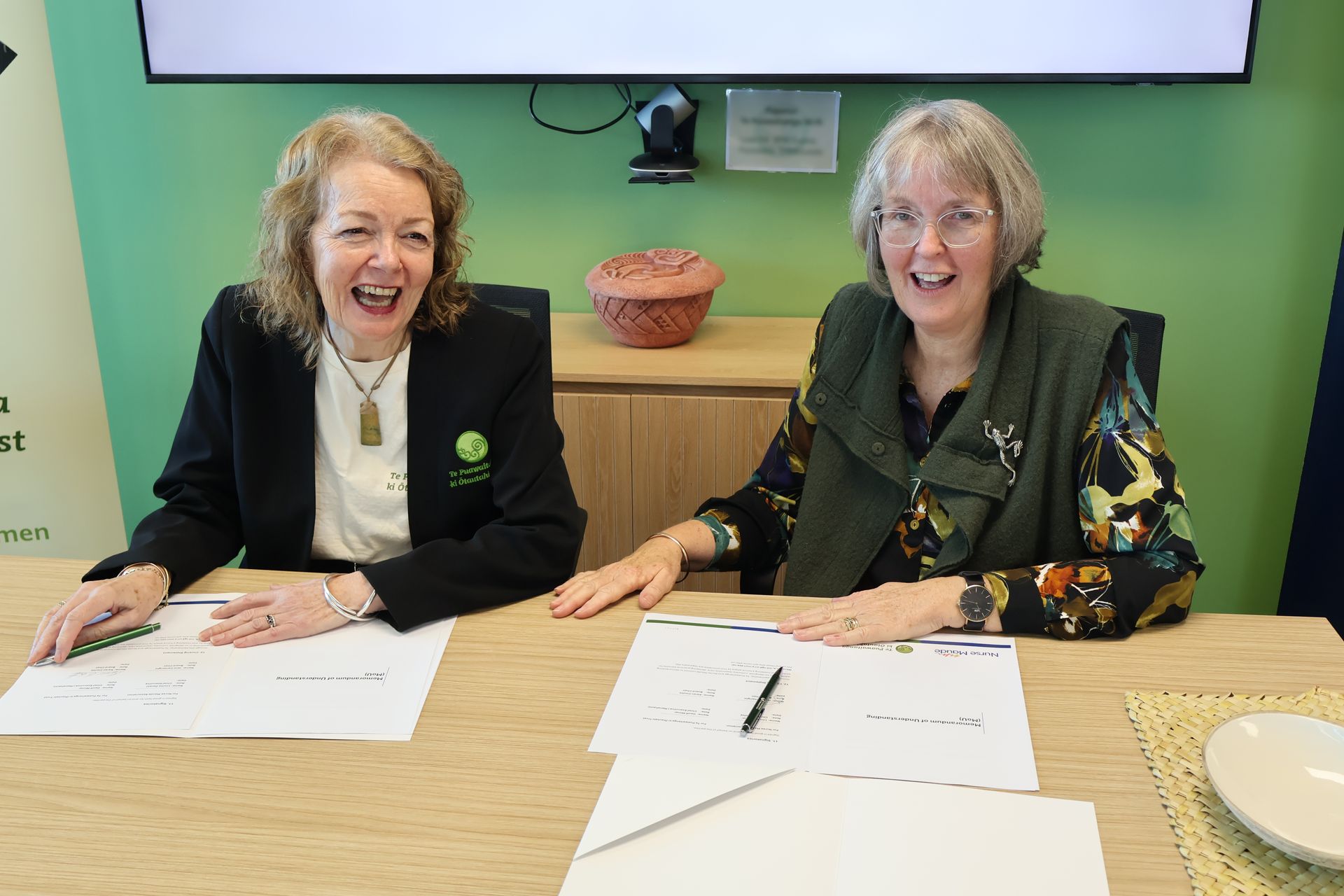 Two women smiling at a table with papers and pens in front of a green wall and screen