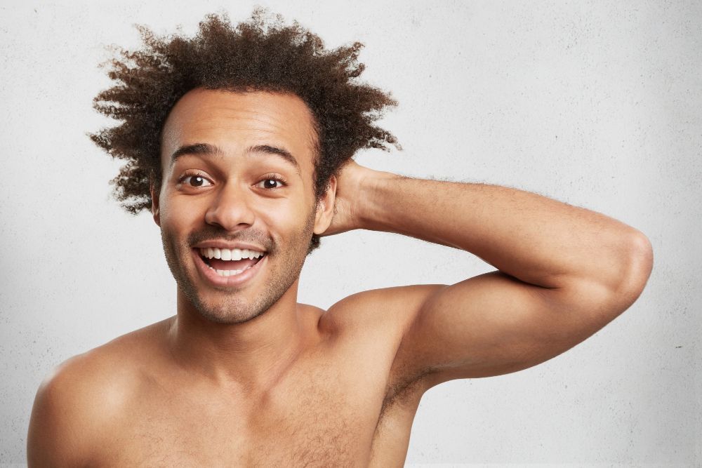 Un homme aux cheveux noirs et bouclés sourit, la main touchant sa tête, sur un fond clair.