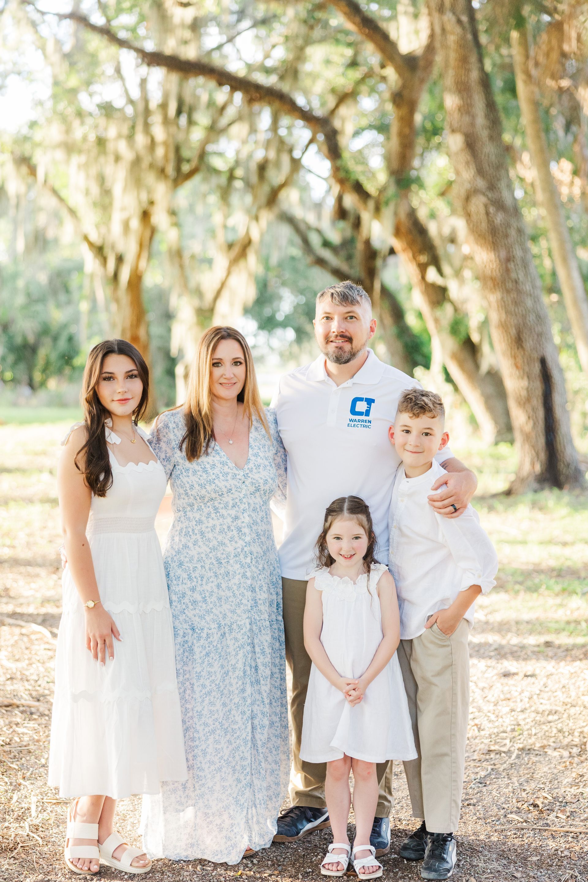 Family of five standing outdoors under trees, posing together in soft sunlight.