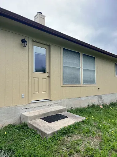 Tan house exterior with front door, window, and concrete steps leading to a small grassy yard