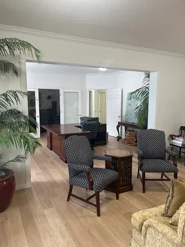 Living room with patterned chairs, round table, sofas, and large potted plants through a wide opening