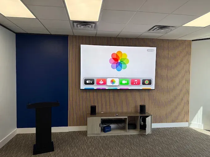Conference room with wall-mounted display, podium, and cabinet beneath a colorful logo on the screen