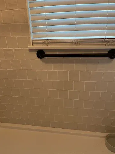 Bathroom wall with beige tile, a window with white blinds, and a black towel bar.