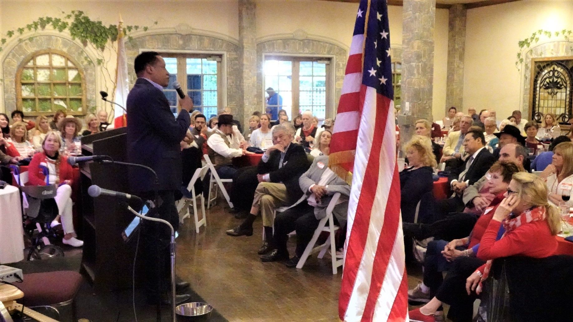 Man speaking at podium, American flag in foreground, audience seated in hall.