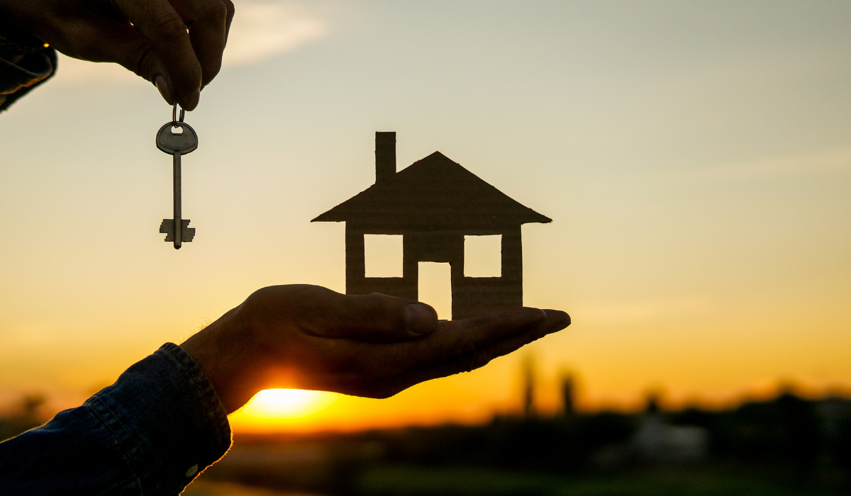 Hand holding a house cutout and key against a sunset background.