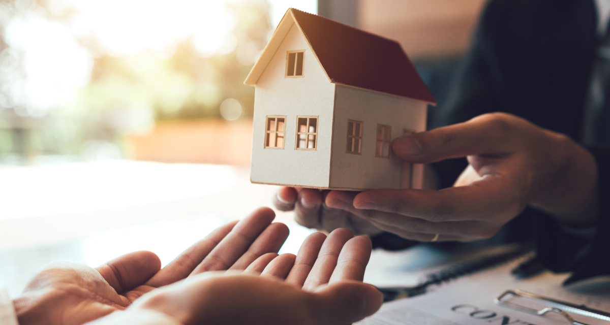 Hands passing a miniature house during a transaction, likely real estate. Sunlight streams in, contract visible.
