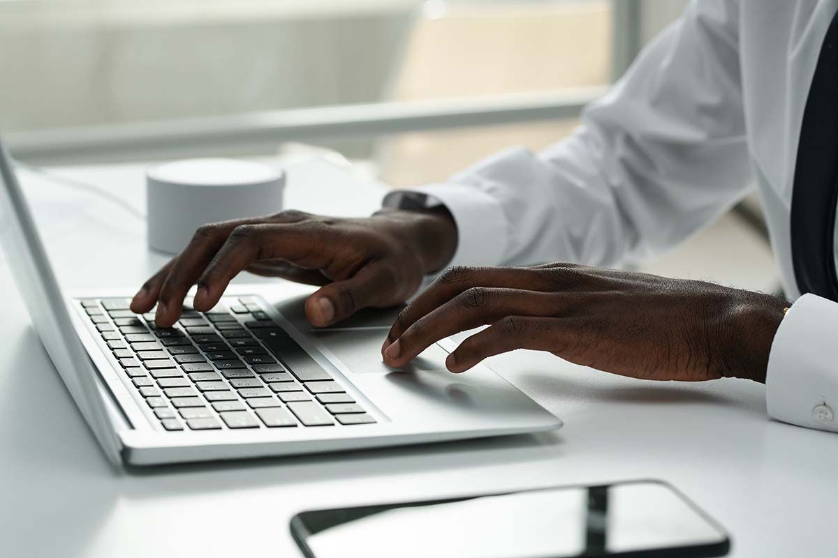 Person typing on a laptop with dark skin, wearing a white shirt and black tie. Phone on the desk.