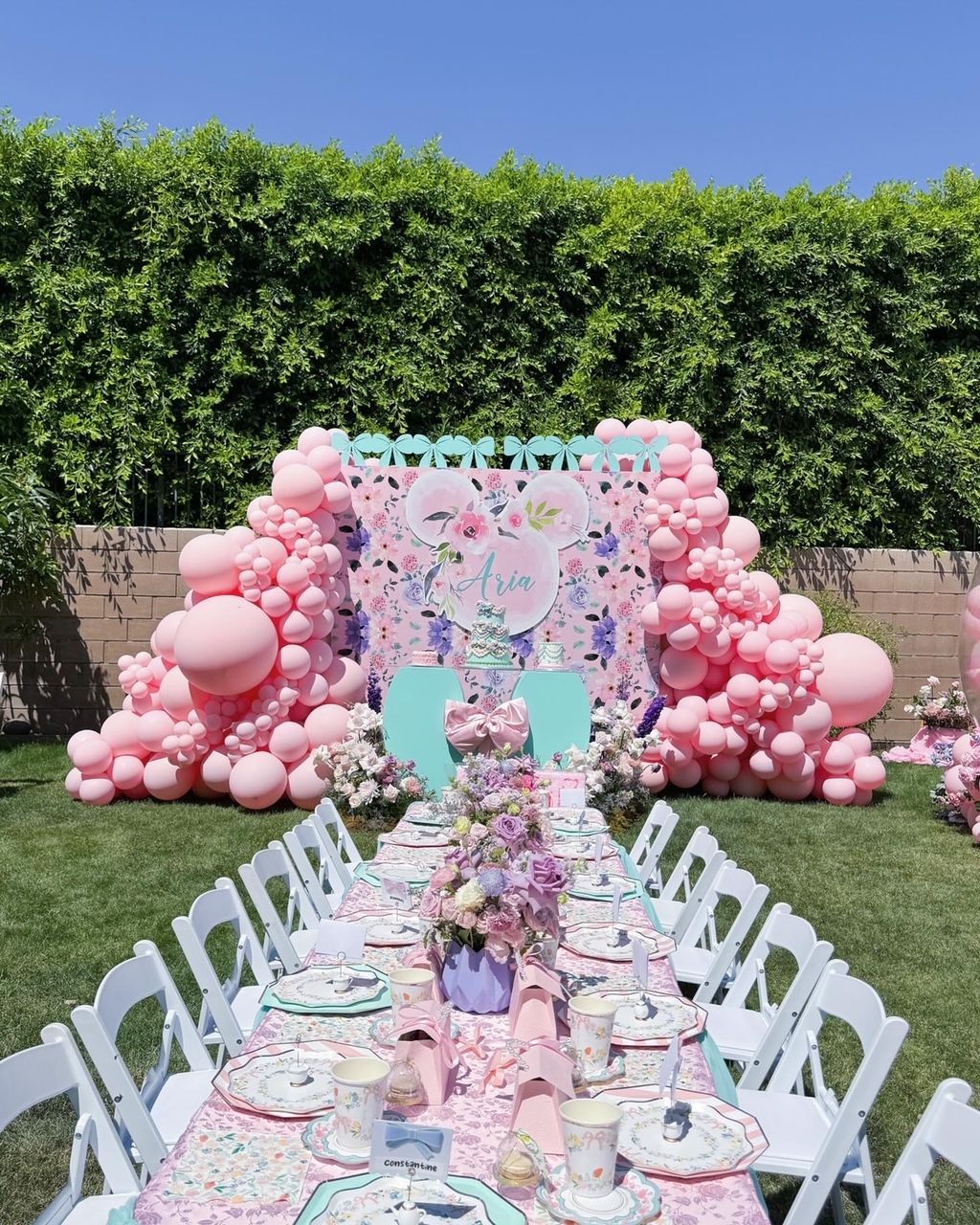 Pastel pink balloon arch around floral backdrop beside a long decorated outdoor party table