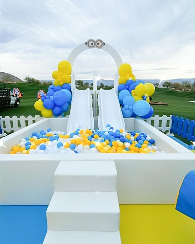 Outdoor party setup with white arch, blue and yellow balloons, and a small white staircase on a green lawn.