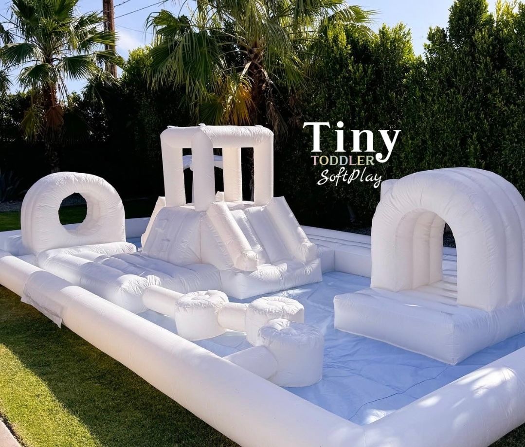 White inflatable play structure with ramps and arches on a lawn, with “Tiny Tots” text in the background