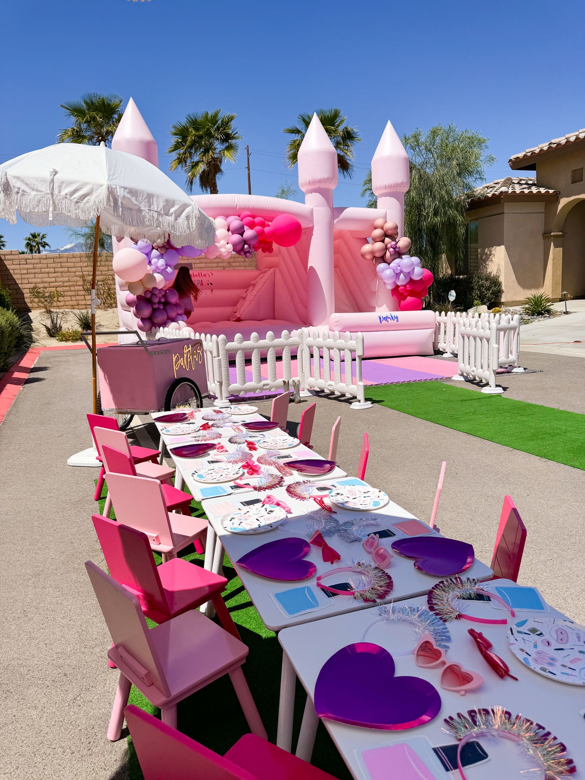 Pink princess-themed party setup with castle bounce house, long decorated table, and white chairs outdoors