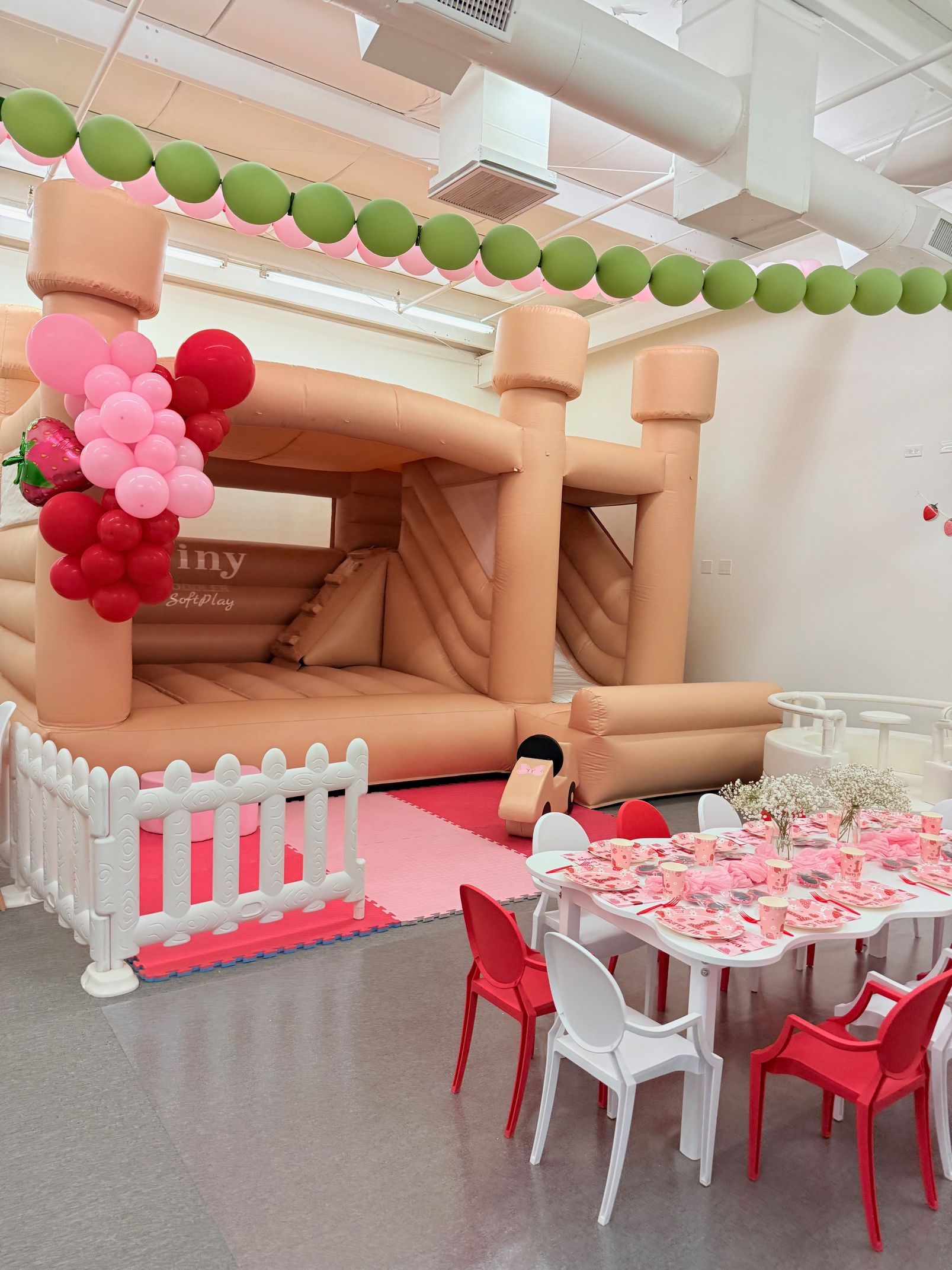 Inflatable bounce house party setup with pink and green balloons, red and white tables, and chairs in an indoor venue