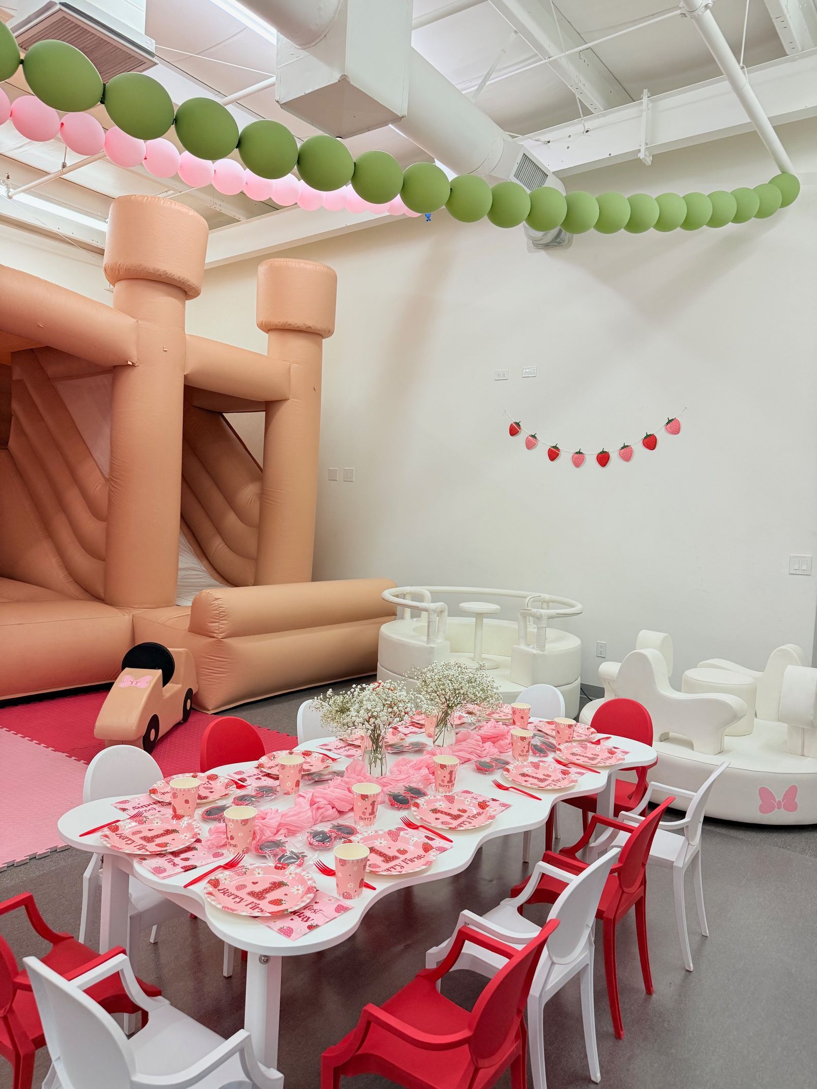 Indoor kids party setup with pink tableware, red chairs, and a large peach inflatable play structure.