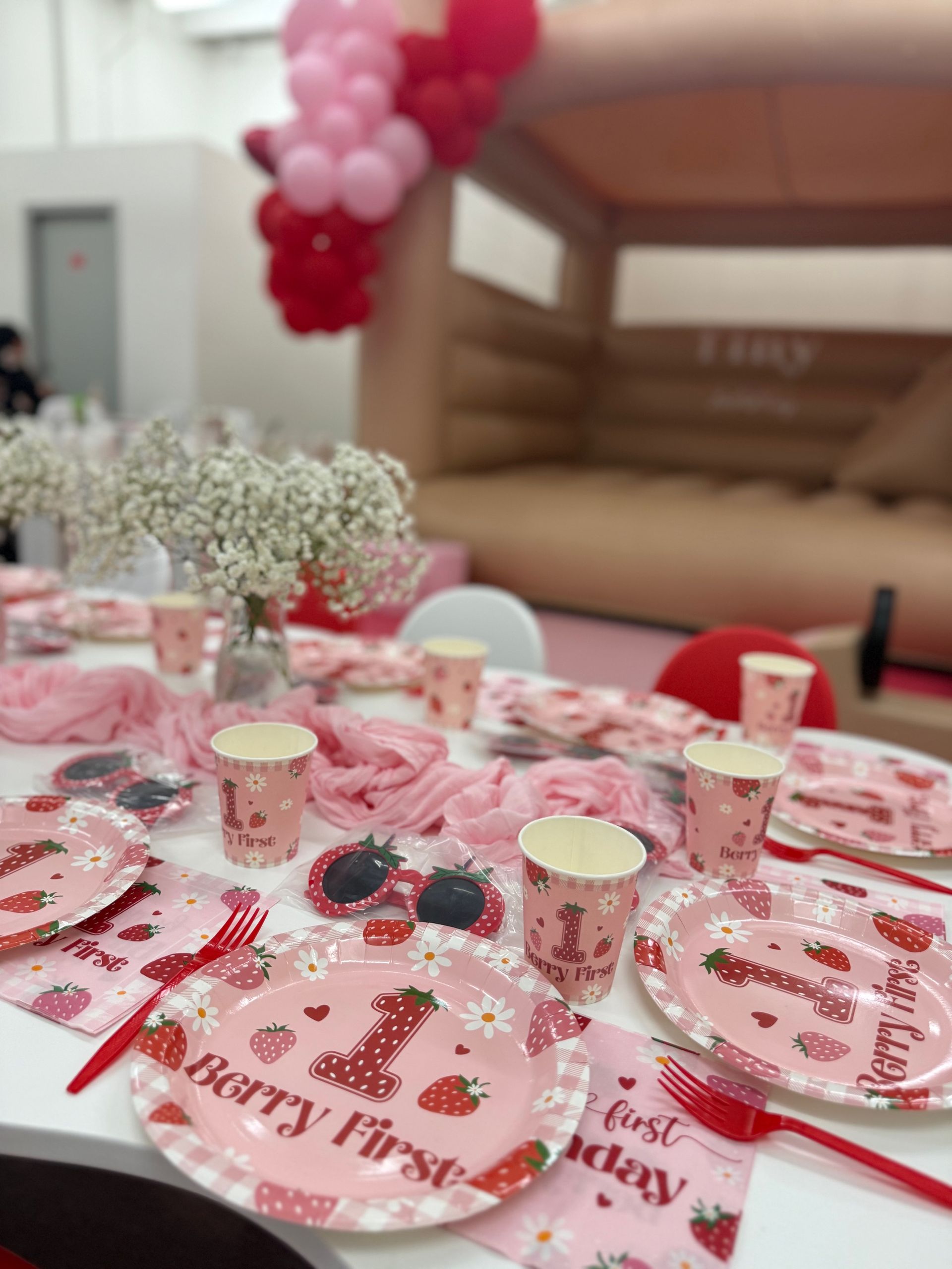 Pink first-birthday table setting with plates, cups, and balloons in a party room