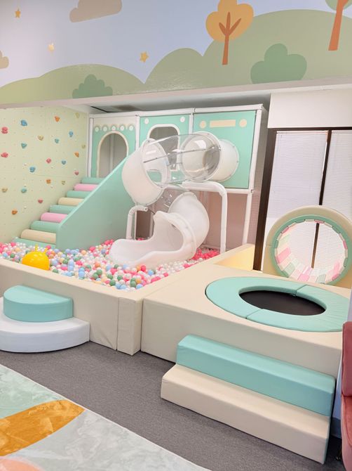 Pastel indoor play area with ball pit, slides, tunnels, and wall-mounted fans beneath a mural
