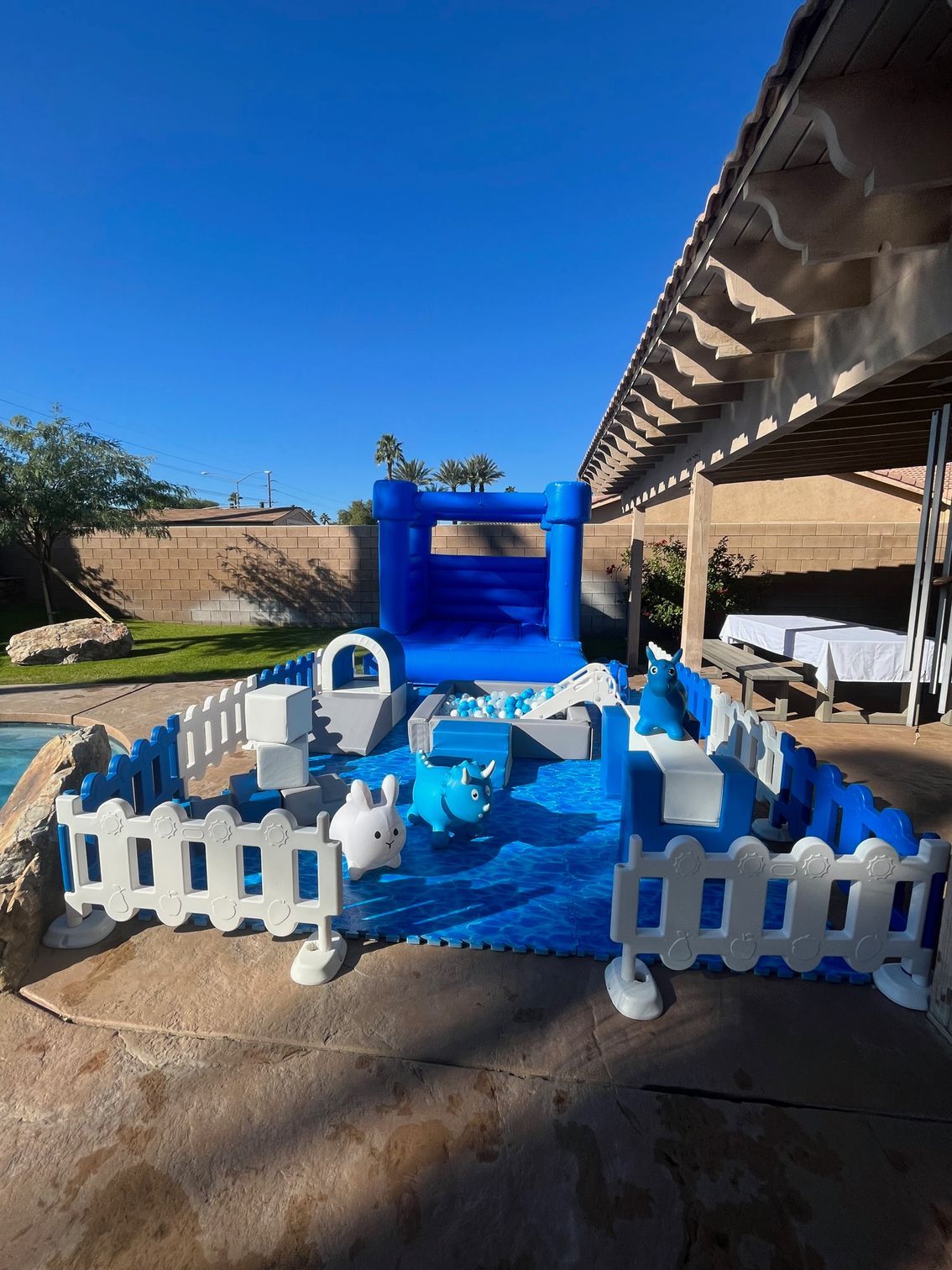 Blue inflatable kiddie pool with white picket fence and toy decorations under a carport in a backyard