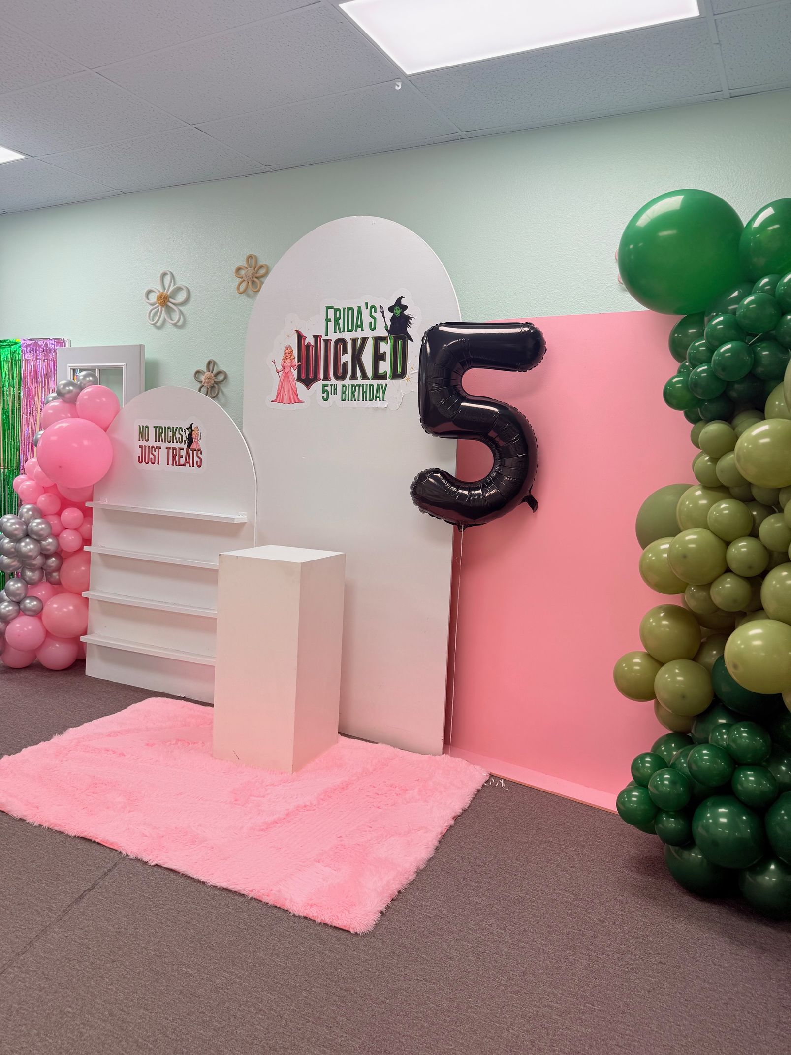 Pink birthday setup with balloon columns, a large black “5,” and white pedestals on a rug in a room