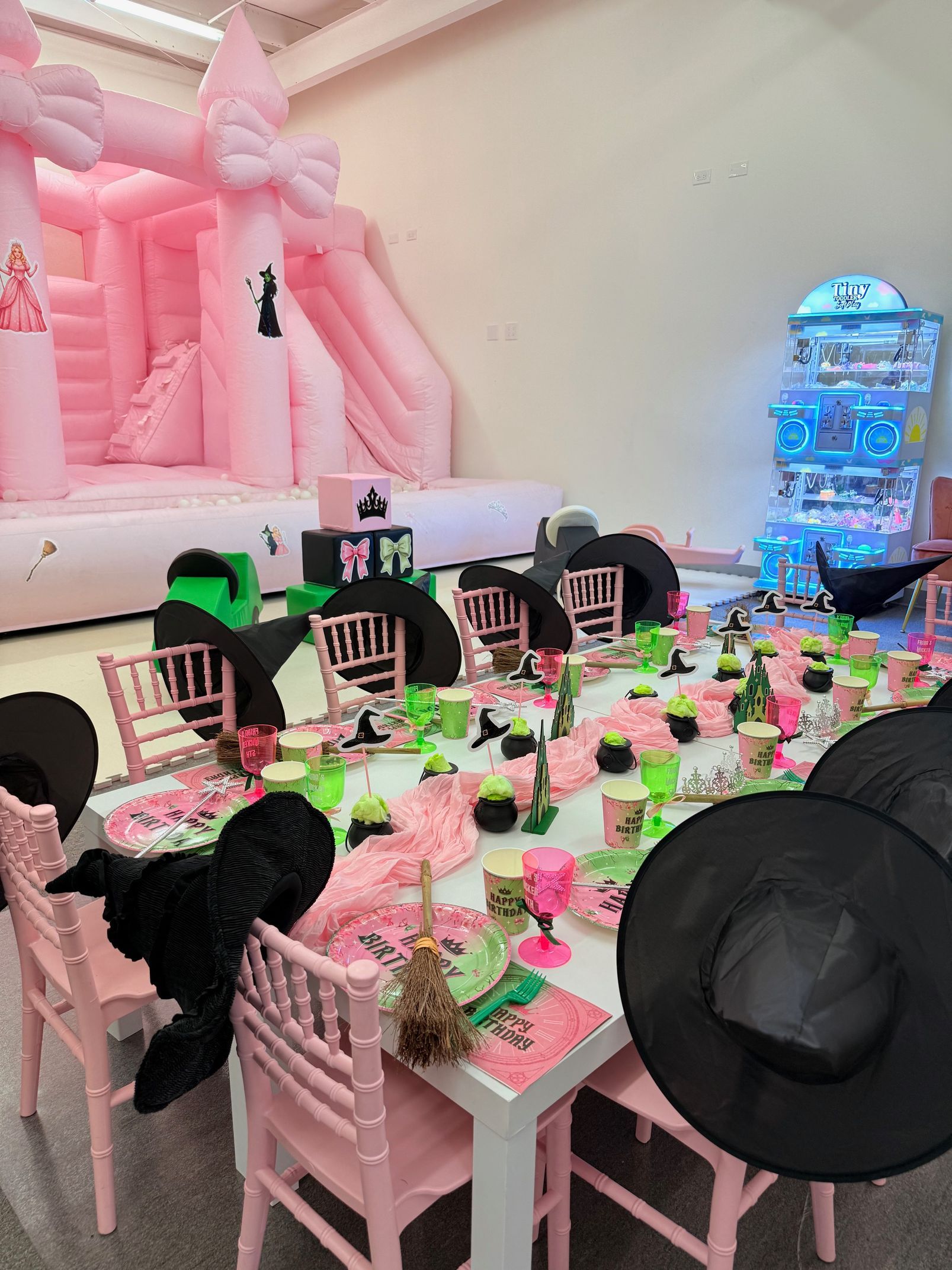 Pink party tables with black chairs in a decorated event room, with balloons and gift boxes along the walls