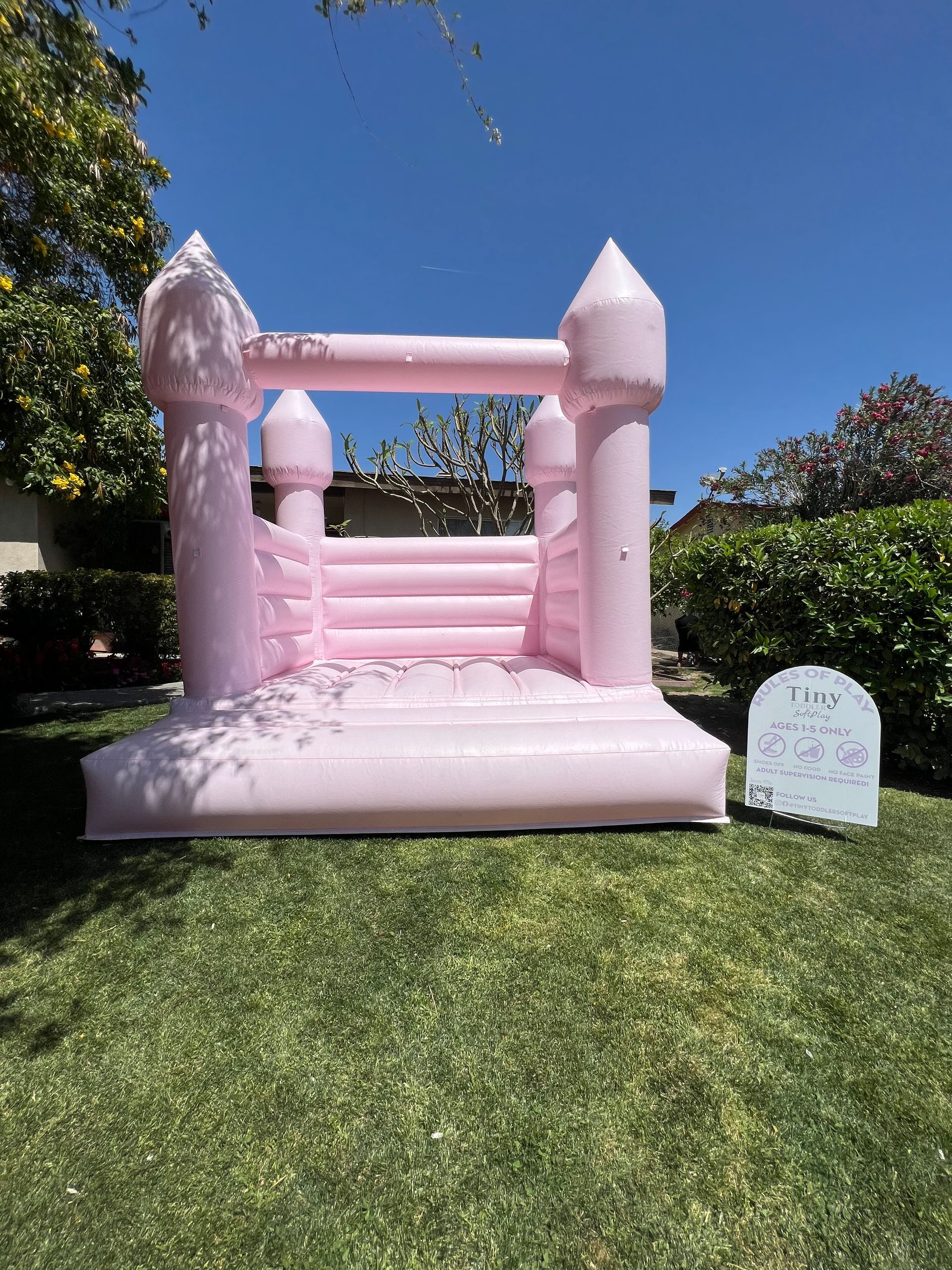 Pink inflatable bounce castle on a lawn in a sunny backyard, with a small sign beside it.